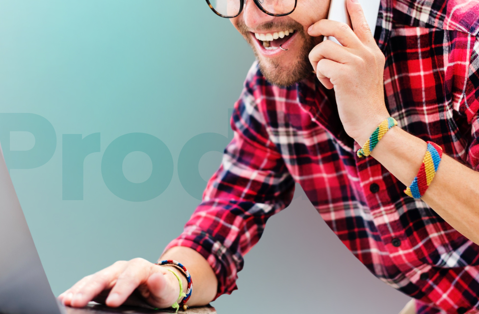 A young man in a flannel shirt is holding a phone and using a laptop